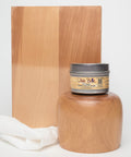 A jar of Dixie Belle wood conditioner is placed in front of a wooden block and a polished wooden object. A white cloth rests beside the jar, all set against a plain white background.