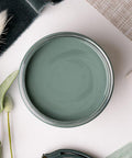 A flat lay of a green-themed arrangement featuring a jar of sage green paint, various shades of green fabric, a green purse, and sprigs of dried foliage on a white surface.