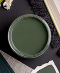 A top view of an open jar of dark sage green paint surrounded by fabric samples, a book, and white flowers on a dark surface. The arrangement suggests a color-coordinated design theme.