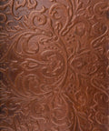 Close-up of a brown leather surface with intricate embossed floral patterns. The design includes swirling leaves and blooms, creating a textured, elegant appearance.