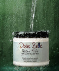 A jar of Dixie Belle Gator Hide is splashed with a stream of water against a green background, demonstrating its water-repellent properties. Water droplets are seen around and on the container.
