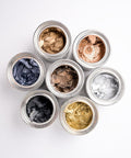 Seven open jars of metallic pigment waxes in gold, silver, bronze, black, and blue shades arranged in a flower-like pattern on a white background.