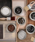 Open cans of wood stain in various colors, with paint brushes resting on the lids and trays, are arranged on a wooden surface alongside stained wood samples in different shades.
