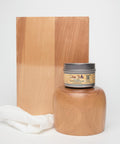 A jar of Dixie Belle wood conditioner is placed in front of a wooden block and a polished wooden object. A white cloth rests beside the jar, all set against a plain white background.