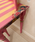 A close-up of a pink painted table with a striped orange-and-pink top, a floral rose-patterned transfer, and turned legs, set against a beige wall and carpet.