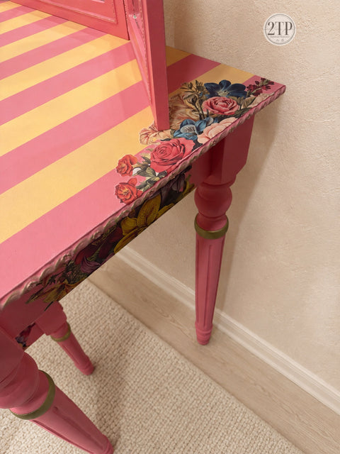 A close-up of a pink painted table with a striped orange-and-pink top, a floral rose-patterned transfer, and turned legs, set against a beige wall and carpet.