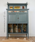 A painted wooden cabinet with a muted blue-gray finish, featuring floral wallpaper panels in the lower storage compartments and a small mirror on the top shelf. The cabinet stands on a wooden floor against a white wall, with a woven rug partially visible in the foreground.