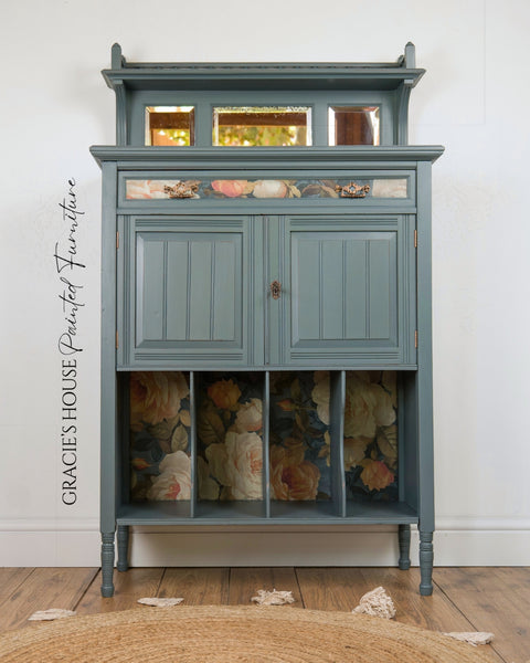 A painted wooden cabinet with a muted blue-gray finish, featuring floral wallpaper panels in the lower storage compartments and a small mirror on the top shelf. The cabinet stands on a wooden floor against a white wall, with a woven rug partially visible in the foreground.