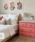 A styled bedroom scene featuring a coral pink three-drawer nightstand with brass knobs next to a bed with white linens and floral accent pillows. The background is a white painted brick wall with three matching floral prints, and a 'Stacey Jordan Designs' logo watermark is in the bottom-left corner.