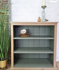 A small bookshelf painted in muted green with a light wood frame, styled with books, decorative objects, and a plant nearby.
