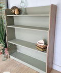 A light green wooden bookshelf with four shelves, holding a glass lamp, decorative items, and a few stacked books. The bookshelf stands on a wooden floor against a white brick wall, with flowers arranged nearby.