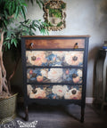 A vintage-style three-drawer chest of drawers with a dark painted frame and floral rose patterns on the drawer fronts, shown in a styled room setting with a potted plant and wall mirror.