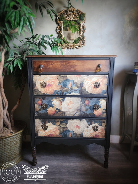 A vintage-style three-drawer chest of drawers with a dark painted frame and floral rose patterns on the drawer fronts, shown in a styled room setting with a potted plant and wall mirror.
