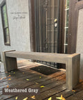 A wooden bench in weathered gray finish placed on a wooden porch. It's positioned near a dark metal door with a mesh screen. Fallen leaves are scattered on the porch. A logo in the corner reads "The Flipped Piece, Shayna Alnwick."