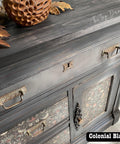 A black wooden dresser with ornate drawer handles and floral patterned drawer fronts. A decorative bowl and autumn leaves rest on top. The words "The Top Drawer Rva" are visible, and "Colonial Black" appears at the bottom.