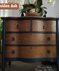 A vintage wooden dresser with four drawers, featuring dark wood accents and elegant metal handles. A vase with orchids and other plants surround it. A label reads "Golden Ash" at the top left corner.