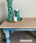 A wooden table with blue legs holds a decorative arrangement of tall, twisted teal candle holders, each with white candles on a brass tray. Nearby, there's a plant in a woven basket. A simple, modern interior design setup.