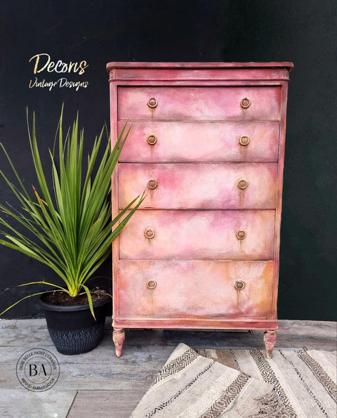 A dresser that has multiple colors of chalk paint from flamingo to peony to fluff all blended beautifully.  there is a plant to the left in a black pot and a rug in front