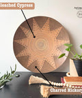 A decorative wooden bowl with a sunburst pattern is displayed on a dark surface. Nearby, a potted plant, decorative beads, a stack of books, and a tassel add to the arrangement. The wall is plain, highlighting the bowl's intricate design.