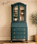 Vintage secretary desk and hutch painted in a rich teal blue finish with ornate carved details, glass cabinet doors, and gold hardware, styled in a bright neutral room.