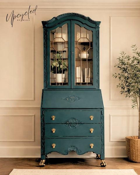 Vintage secretary desk and hutch painted in a rich teal blue finish with ornate carved details, glass cabinet doors, and gold hardware, styled in a bright neutral room.