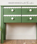 Green side table with white trim and drawers next to a yellow vase on a wooden floor.