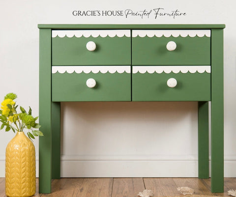 Green side table with white trim and drawers next to a yellow vase on a wooden floor.