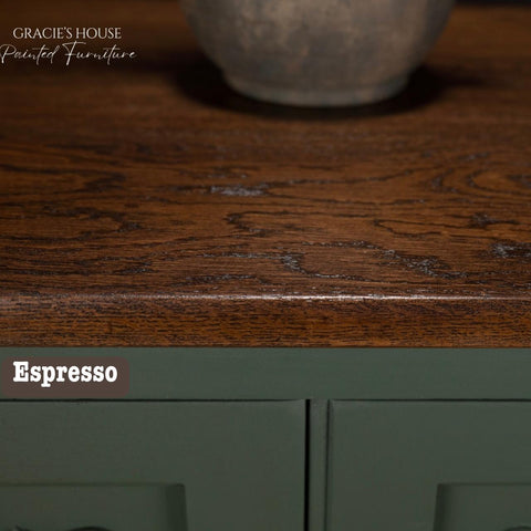 Close-up of a wooden dresser top with a rich, dark espresso finish and subtle texture. The top shows natural wood grain, contrasting with a deep green-painted front panel below. A round ceramic pot is partially visible on the top.