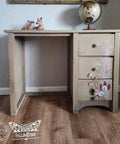 A vintage wooden desk with three drawers adorned with painted floral designs. A small globe and two decorative deer figurines sit on top. A textured wall and framed art are in the background, with logos in the bottom left corner.