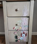 A wooden dresser with three drawers. The front showcases a floral design with various flowers in soft colors. A logo featuring the text "FallinReine" and a circular emblem are on the bottom left. The dresser stands on a wooden floor.