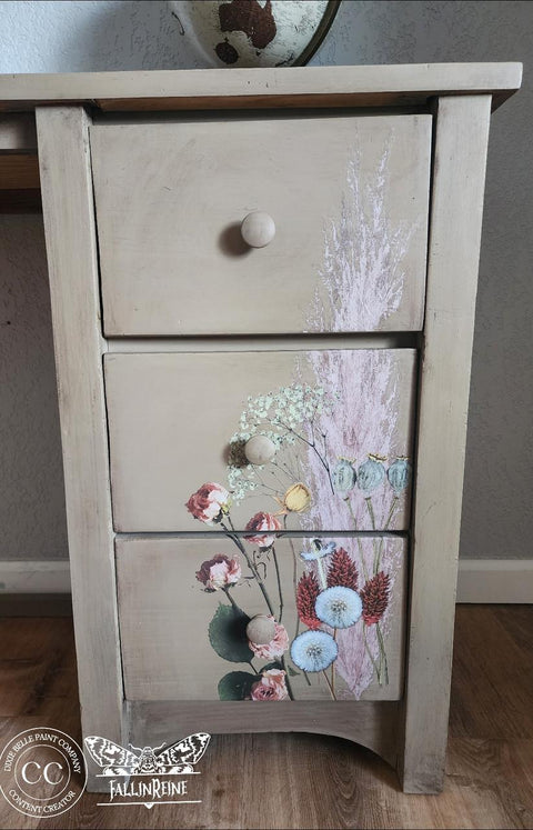 A wooden dresser with three drawers. The front showcases a floral design with various flowers in soft colors. A logo featuring the text "FallinReine" and a circular emblem are on the bottom left. The dresser stands on a wooden floor.