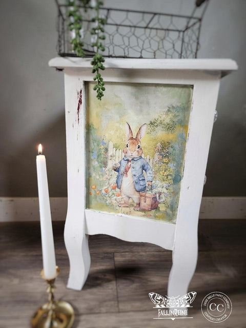 White wooden nightstand with a decoupage of a rabbit in a blue jacket on the front panel. A lit candle in a holder stands nearby. The scene has a whimsical, garden setting, and a small plant is on the tabletop. A watermark is visible at the bottom.