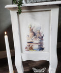 A white painted nightstand with a vintage illustration of a rabbit surrounded by books on the side. A lit candle stands beside the table. There are green plants on top of the table.