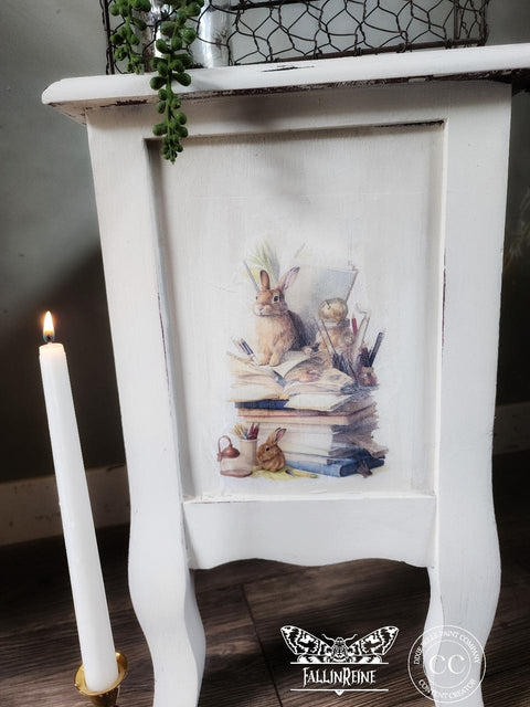 A white painted nightstand with a vintage illustration of a rabbit surrounded by books on the side. A lit candle stands beside the table. There are green plants on top of the table.