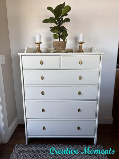 White dresser with six drawers and gold knobs, topped with a potted plant and two white candles in gold holders, standing against a light-colored wall with "Creative Moments" text in the bottom right corner.