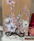 A wooden plaque with delicate floral artwork featuring pink and white flowers, with white leaves and small berries. It stands on a black holder, placed on a soft white surface. Text reads "Reimagined by Patty" in elegant script.