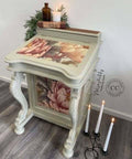 Vintage-style desk with floral patterns on the top and side. Books and lit candles are on the desk, with a decorative metal candle holder on the floor. The desk is set against a white wall on a wooden floor.