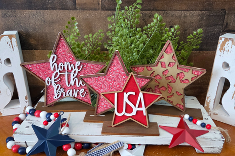 Decorative stars with 'home of the brave' and 'USA' text on a wooden surface with plants in the background.
