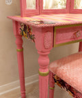 A pink vanity table with a floral and striped patterned top, matching pink pedestal legs with gold trim, and a coordinating pink upholstered bench in a styled interior setting.