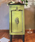 A small, ornate green cabinet with black wax accents stands on a wooden floor. An open book and a plant adorn the top. A paper is on the floor nearby.