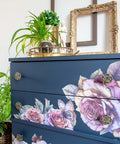 A navy blue three-drawer dresser decorated with large pink and purple floral prints and butterfly motifs, styled in a home setting with a gold mirror, potted plants, and decorative knobs.