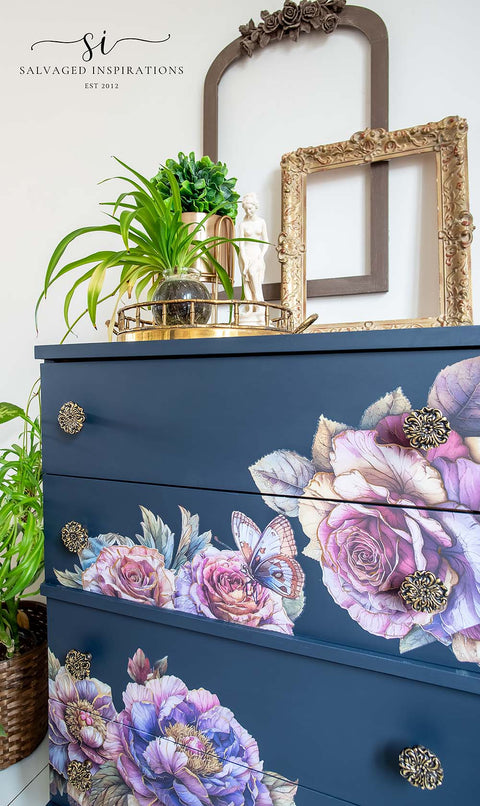 A navy blue three-drawer dresser decorated with large pink and purple floral prints and butterfly motifs, styled in a home setting with a gold mirror, potted plants, and decorative knobs.