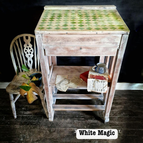 Vintage desk with a worn wood finish, featuring a green diamond pattern on top. A shelf holds an antique book and newspaper. A rustic chair sits beside it, adorned with leaves. A small, decorative cup is placed on the shelf. Text reads "White Magic."