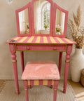 A pink vanity table with a three-panel mirror, striped yellow and pink tabletop, and a matching pink plush stool, styled in a warm neutral room with dried grass in a vase.