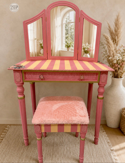 A pink vanity table with a three-panel mirror, striped yellow and pink tabletop, and a matching pink plush stool, styled in a warm neutral room with dried grass in a vase.