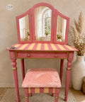 A pink vanity table with a three-panel mirror, striped orange and pink tabletop, and a matching pink plush stool, styled in a warm neutral room with dried grass in a vase.