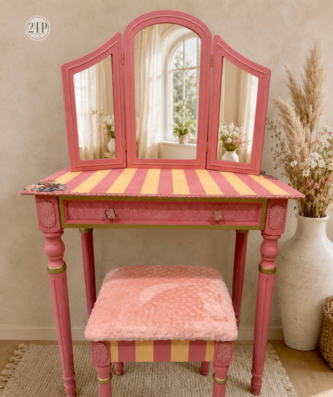 A pink vanity table with a three-panel mirror, striped orange and pink tabletop, and a matching pink plush stool, styled in a warm neutral room with dried grass in a vase.
