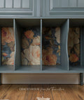 A painted wooden storage bench or cabinet with four open cubbies, featuring a muted blue-gray finish and floral decoupage inside the compartments. The floor is a medium brown wood, and a small piece of rope lies on the floor in the foreground.