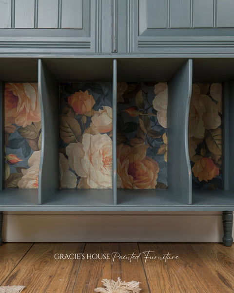 A painted wooden storage bench or cabinet with four open cubbies, featuring a muted blue-gray finish and floral decoupage inside the compartments. The floor is a medium brown wood, and a small piece of rope lies on the floor in the foreground.