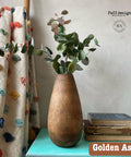 A wooden vase with eucalyptus branches is on a turquoise table next to stacked books. A colorful, textured curtain hangs beside it. The text reads "Faff designs" and "Golden Ash."